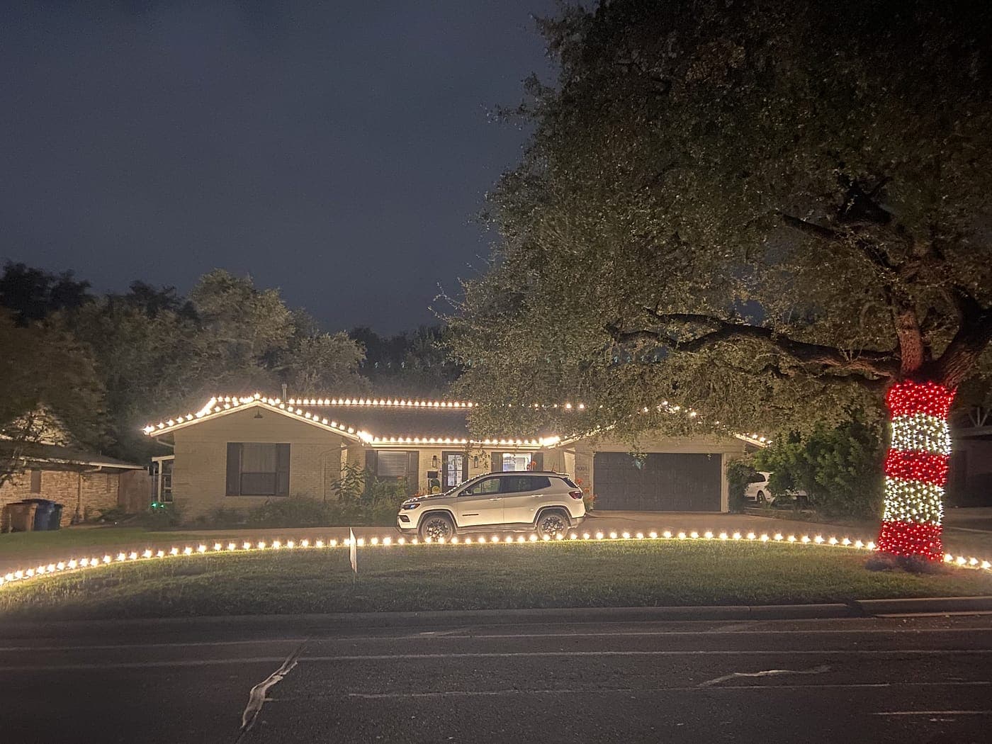 A single-story home with a bulb roofline, lights along the full length of the lawn edge, and a tree wrapped candy-cane style